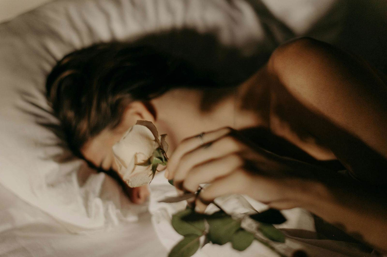 Woman laying on bed holding white rose with her head buried in pillow