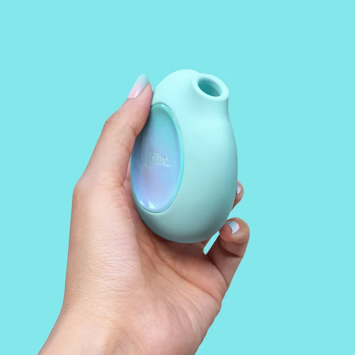 A hand with light pink manicured nails holds a small, rounded, mint green Biird Lumii Clitoral Suction Stimulator silicone adult toy with a transparent circular window showing a blue-purple interior, set against a solid light blue background.