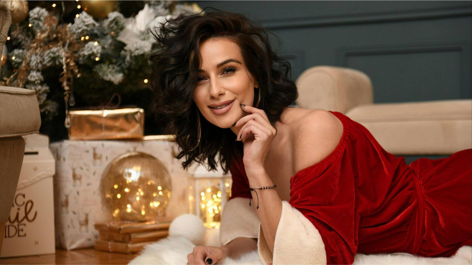 Woman with wavy brown hair on the floor in a Santa Claus outfit looking flirty and seductive in front of Christmas tree and holiday gifts.