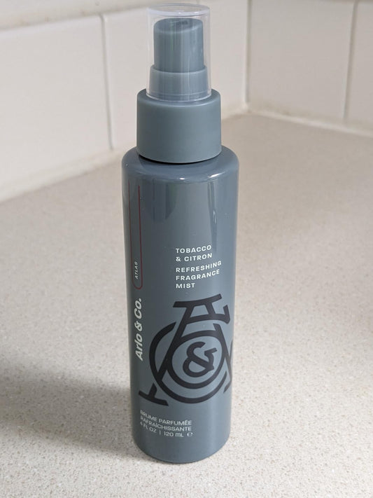 A bottle of Pure Romance Arlo & Co Refreshing Fragrance Mist in Tobacco & Citron scent, standing on a light countertop against a tiled backsplash.