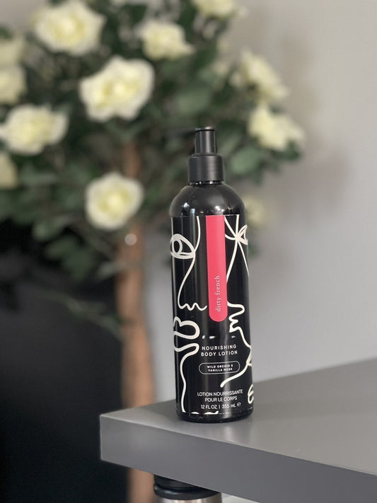 Pump bottle of Pure Romance 'dirty french' Nourishing Body Lotion. The bottle is black with a black pump, decorated with white minimalist line art of faces and a vertical pink label with flowers in the background.