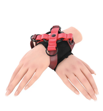 A red and black wrist harness by Sportsheets Sex Mischief brand with black metal rings, displayed on a mannequin's two hands against a plain white background.