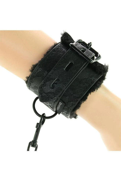 A black lace Sportssheets fur-lined handcuff with fur trim, a blck-tone buckle, and a dangling black clip, shown worn on a person's wrist against a white background.