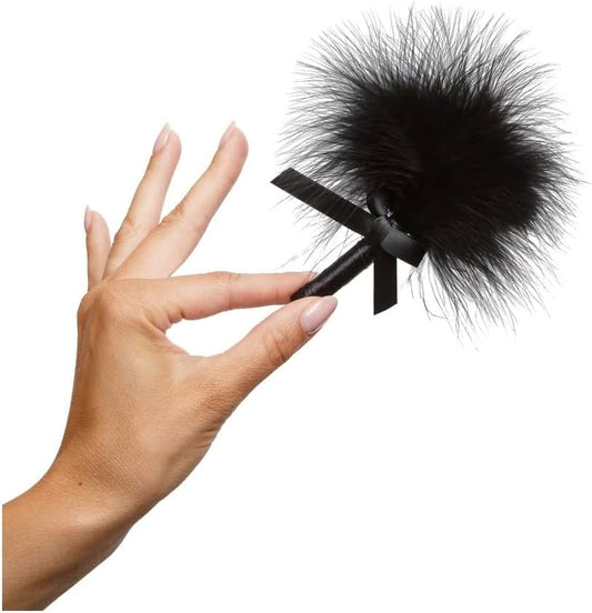A hand holding a black feather tickler applicator featuring a black ribbon handle against a plain white background that is included in the package with Kama Sutra Honey Dust Raspberry Kiss.