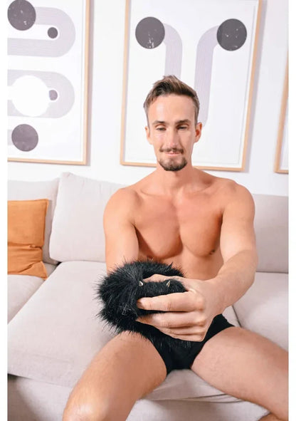 Man sitting on a couch holding a Sportsheets black fur spiked sensory mitt with abstract wall art in the background.
