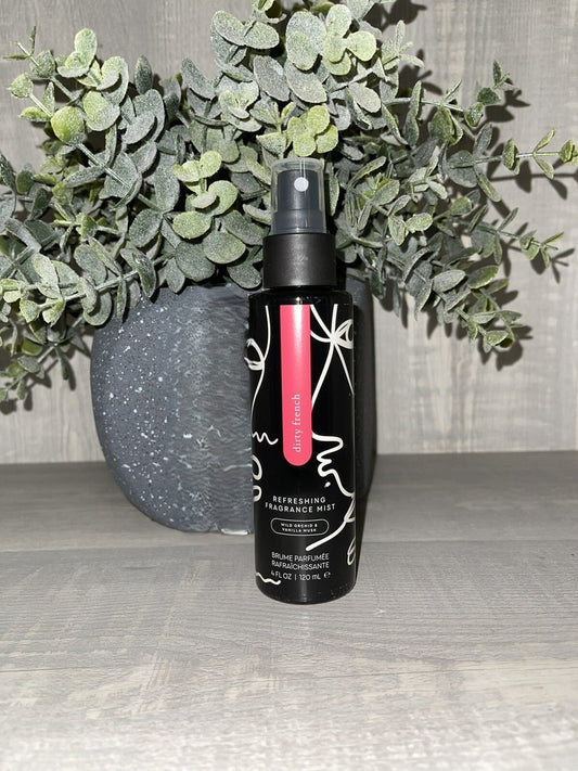 A bottle of Pure Romance Dirty French Refreshing Fragrance Mist in black bottle with a pink label, placed on a gray wooden surface in front of a gray speckled vase filled with green eucalyptus leaves.