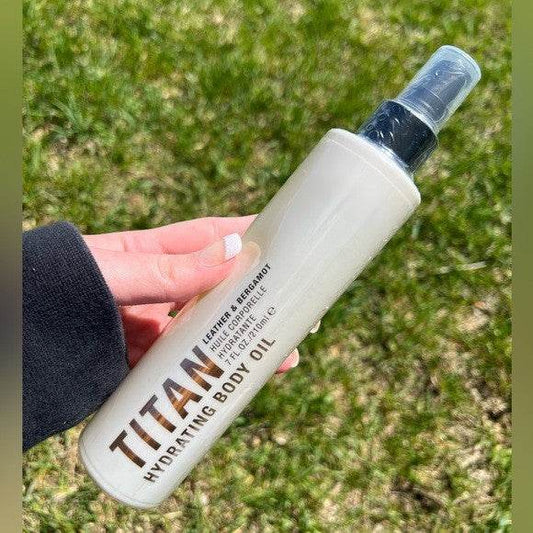 Hand holding a bottle of Pure Romance Titan Hydrating leather bergamot Body Oil against a grassy background