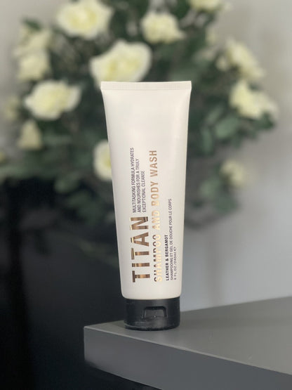 A white squeeze tube of Pure Romance Titan Leather & Bergamot Body Wash with a black cap, standing on a gray surface with a blurred background of white flowers.