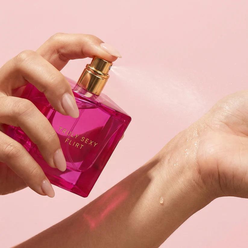 Hand applying pink 'Truly Sexy Flirt' perfume to arm against a pink background