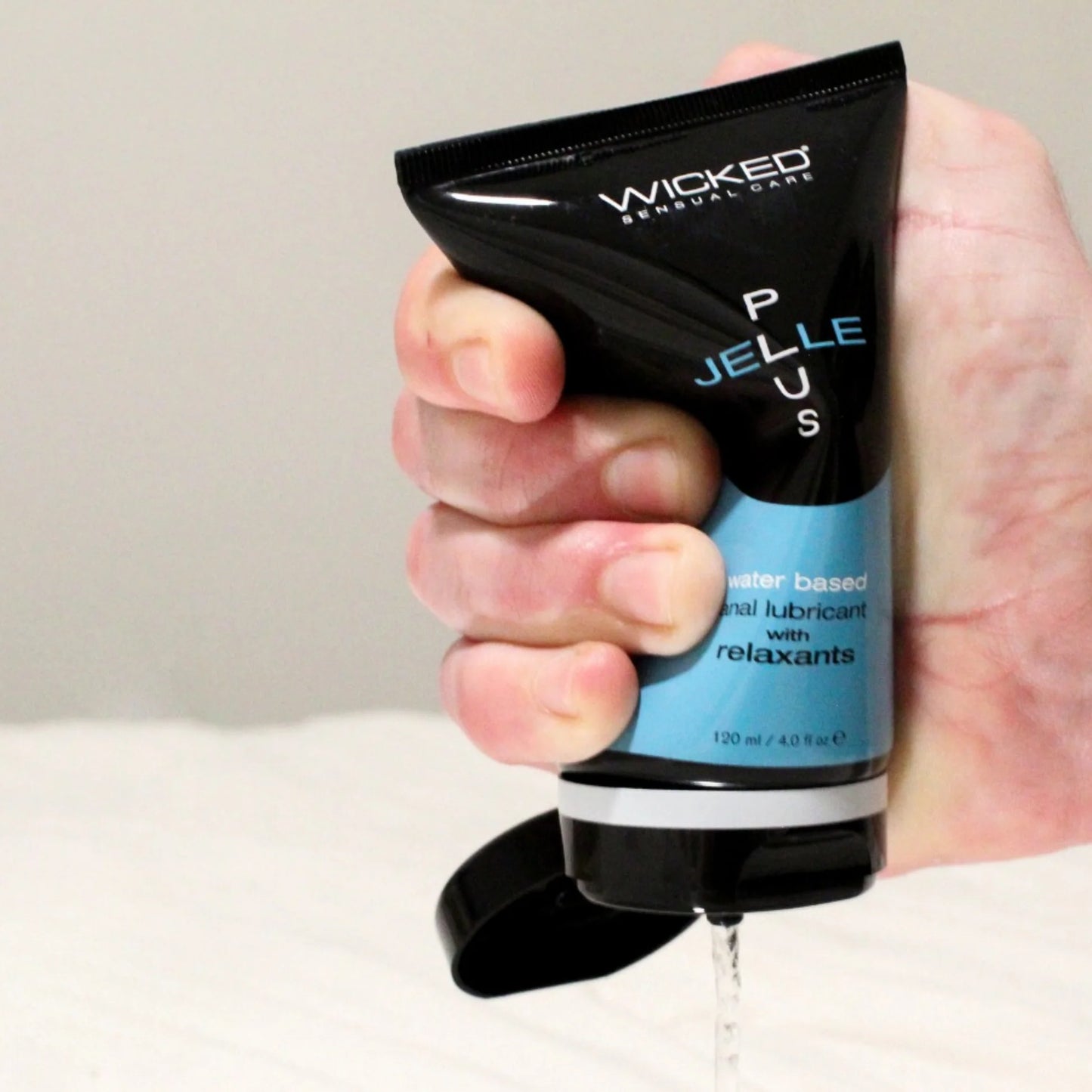 Hand squeezing a Wicked Jelle Plus Water Based Anal Lubricant with relaxants with a neutral background